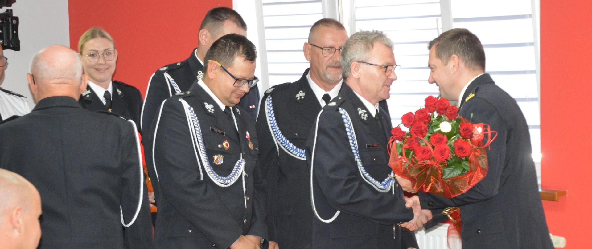 Świetlica komendy. Przedstawiciele władz ZOSP RP składają podziękowania i zyczenia komendantowi powiatowemu. Komendant trzyma w rękach otrzymany bukiet czerwonych róż.