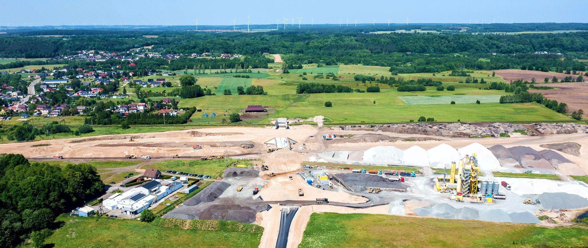 Widok z lotu ptaka na plac budowy. Widoczne są hałdy piasku lub żwiru oraz maszyny budowlane. W okolicy znajdują się pola, las, łąki oraz osiedle z wieloma domami w oddali. 
