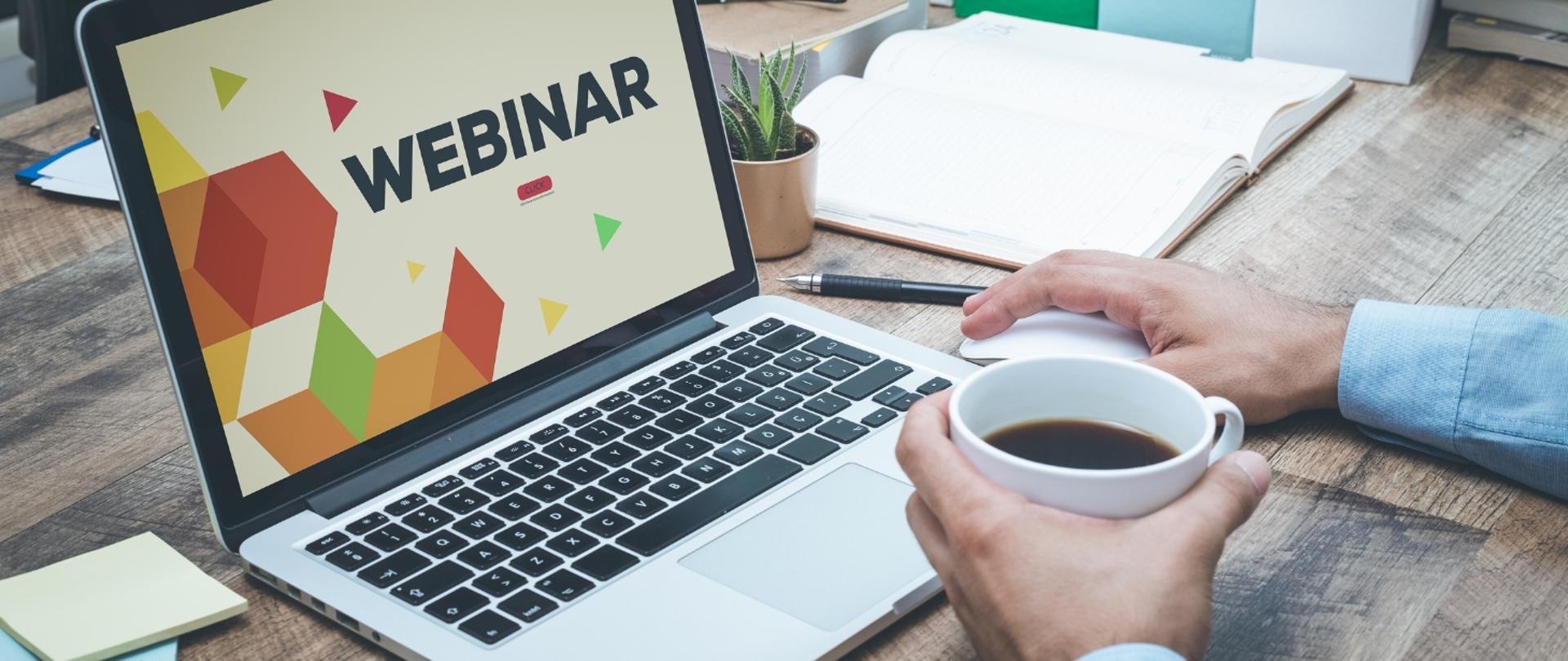 Na zdjęciu widnieje laptop z informacją o webinarze oraz widoczne są dłonie mężczyzny trzymające kubek, w którym znajduje się kawa.