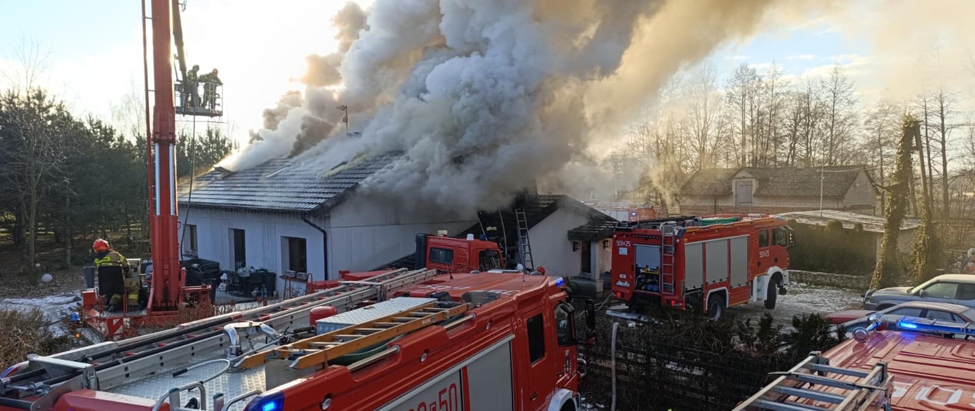 pali się dom jednorodzinny cały dach jest zadymiony i dużo dymu unosi sie do góry obok domu stoją czteery wozy strażacki 