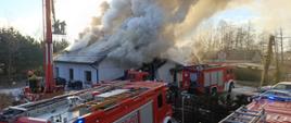 pali się dom jednorodzinny cały dach jest zadymiony i dużo dymu unosi sie do góry obok domu stoją czteery wozy strażacki 