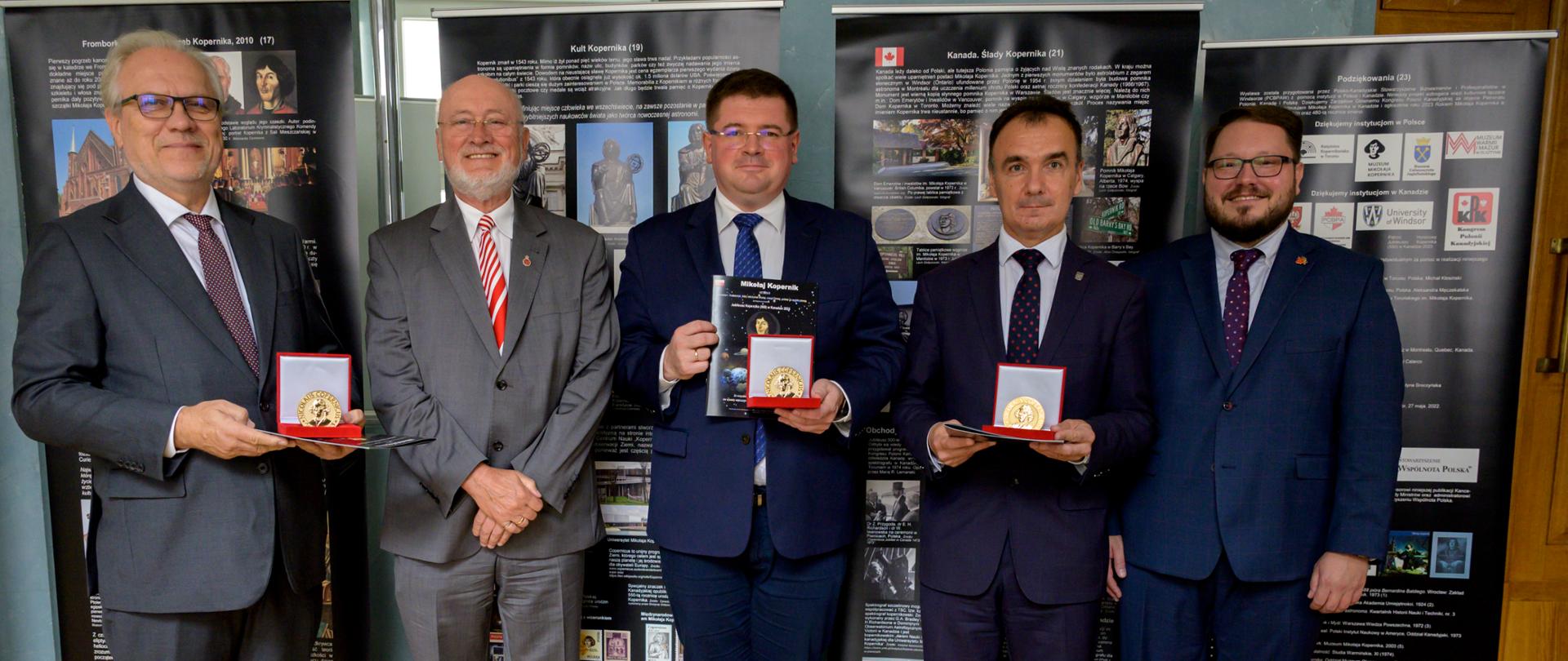 Pod ścianą przy której stoi szereg banerów z napisami i grafikami stoi pięciu mężczyzn w garniturach, trzej trzymają małe pudełeczka w których widać medale.