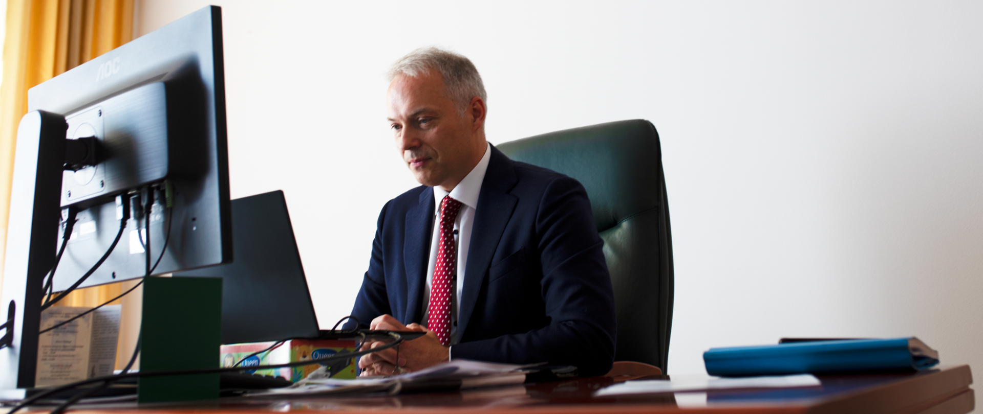 Jacek Żalek w gabinecie siedzi przy biurku z komputerem. Za nim na ścianie godło Polski.