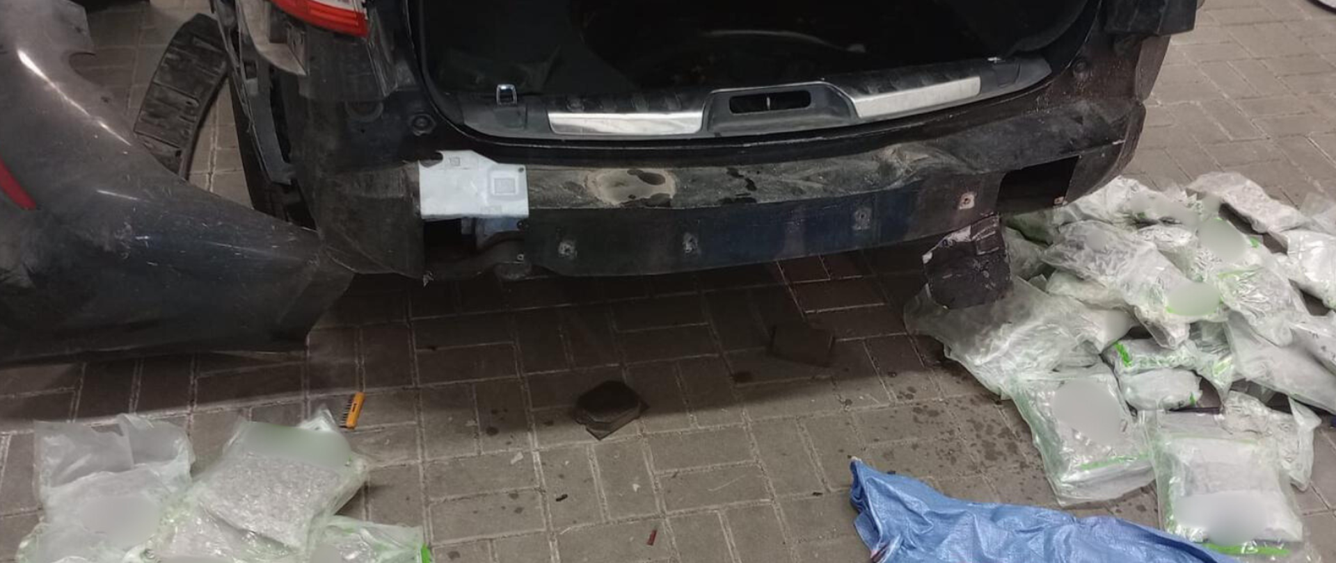 Zdjęty tylni zderzak samochodu. W progach samochodu widoczne opakowania z marihuaną. Przed samochodem wyłożone paczki z narkotykami.