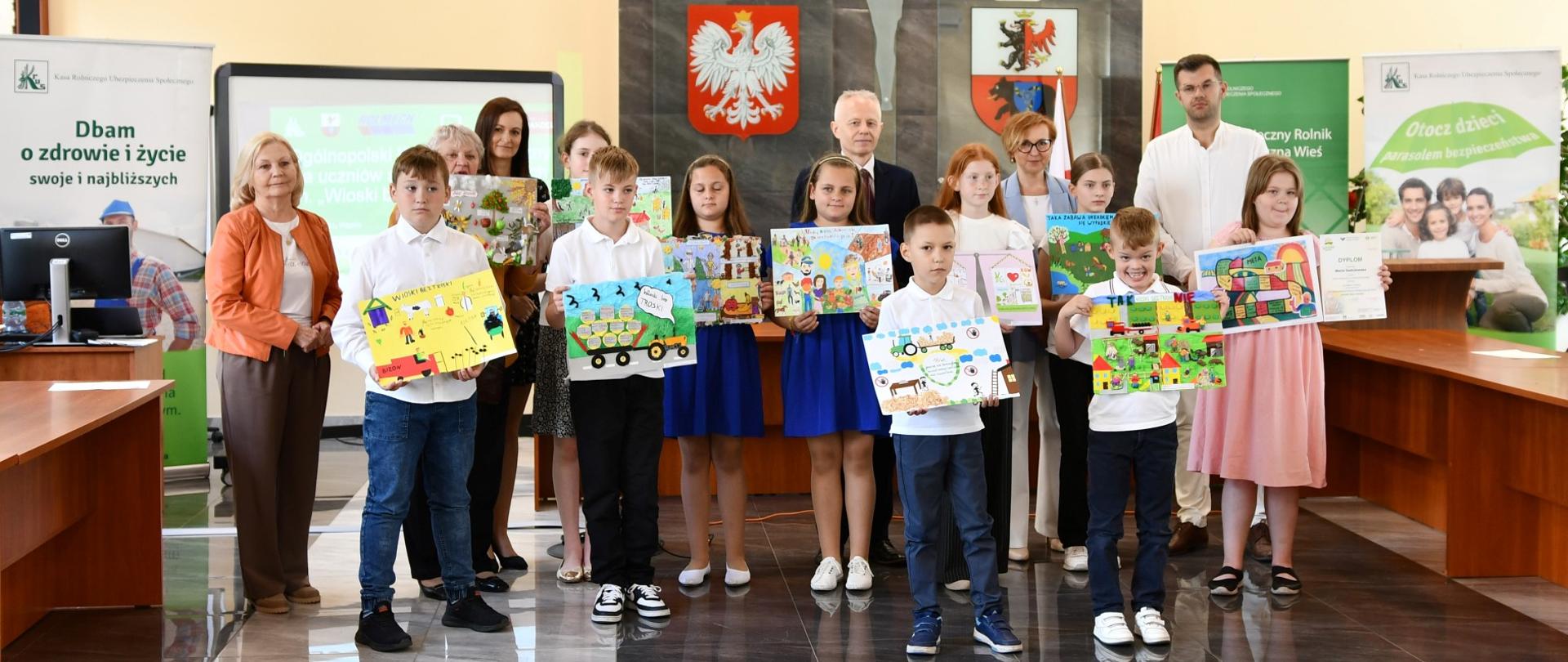 W reprezentacyjnej sali na tle rollupów z logo KRUS pozuje do zdjęcia sześć dziewczynek i czterech chłopców. Dzieci trzymają w dłoniach swoje prace plastyczne, za nimi stoją cztery kobiety i dwóch mężczyzn.