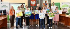 W reprezentacyjnej sali na tle rollupów z logo KRUS pozuje do zdjęcia sześć dziewczynek i czterech chłopców. Dzieci trzymają w dłoniach swoje prace plastyczne, za nimi stoją cztery kobiety i dwóch mężczyzn.