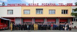 Grupowe zdjęcie przed budynkiem Komendy Powiatowej Państwowej Straży Pożarnej z dużym napisem na dachu 'PAŃSTWOWA STRAŻ POŻARNA TEL. 998'. Przed remizą stoi kilkadziesiąt osób: strażacy w mundurach galowych i bojowych, przedstawiciele władz, zaproszeni goście oraz rodziny z dziećmi. Po lewej stronie kadru widać czerwony pojazd specjalistyczny z wysuniętym zadaszeniem.