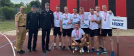 Grupowe zdjęcie reprezentacji województwa łódzkiego w sporcie pożarniczym, wykonane na tle wspinalni Centralnej Szkoły PSP w Częstochowie. Zawodnicy w białych koszulkach z medalami na szyjach, stoją obok trzech funkcjonariuszy w umundurowaniu wyjściowym. Przed nimi widoczne są puchary i dyplomy za osiągnięcia podczas 41. Międzynarodowych Mistrzostw Polski w Sporcie Pożarniczym. Drużyna trzyma tabliczkę z napisem „Województwo Łódzkie”.