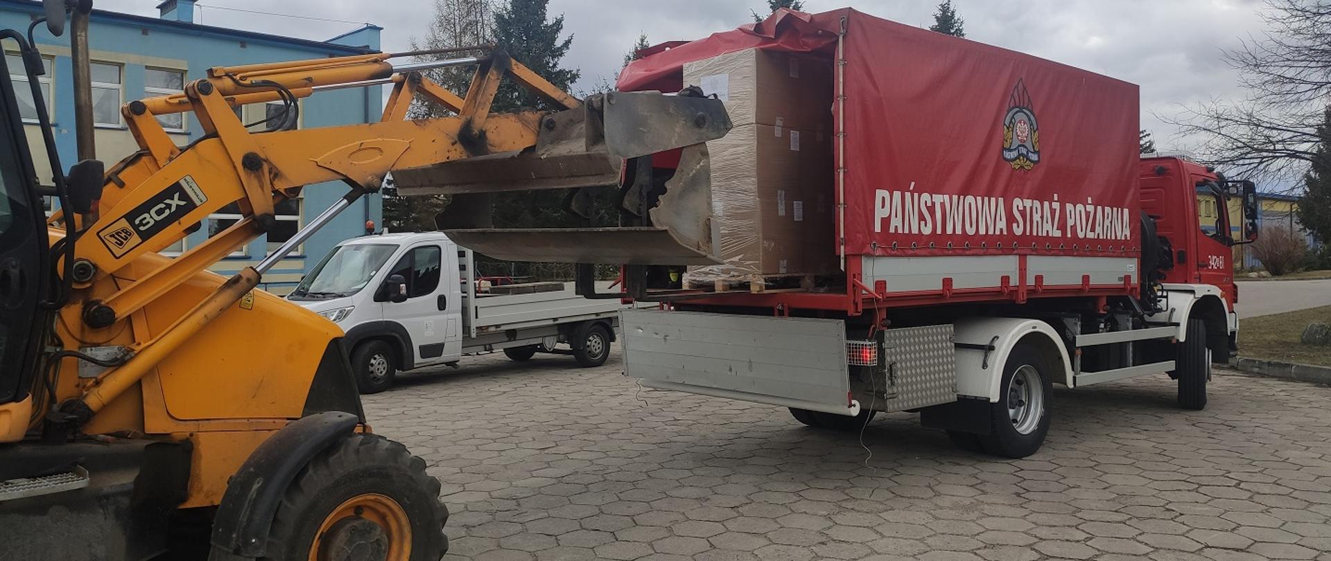 Zdjęcie obrazuje załadunek rzeczy do samochodu strażackiego przy użyciu ładowarki w tle samochód i budynek