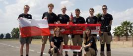 Na betonowym pasie startowym stoi sześciu mężczyzn w czarnych koszulkach, trzymają czerwony bezzałogowy samolot, dwóch z lewej trzyma polską flagę, przed nimi klęczą dwie dziewczyny, w tle palmy.