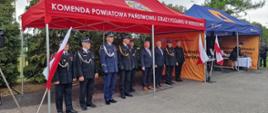 Na zdjęciu widać oficjalne wydarzenie organizowane przez straż pożarną. Uczestnicy stoją pod dwoma namiotami wystawowymi ustawionymi na placu. Czerwony namiot ma napis „Komenda Powiatowa Państwowej Straży Pożarnej w Wieruszowie” oraz emblemat straży pożarnej. Pod nim stoją strażacy w galowych mundurach, funkcjonariusz Policji w niebieskim mundurze oraz przedstawiciele samorządu w garniturach. Wszyscy ustawieni są w szeregu podczas uroczystości o formalnym charakterze. Obok znajduje się niebieski namiot z symbolami ochrony ludności. Na pomarańczowych ściankach widnieje napis „Ochrona ludności i obrona cywilna” oraz logo Ministerstwa Spraw Wewnętrznych i Administracji. Przed namiotami ustawiono biało-czerwone flagi Polski. W tle widoczne są drzewa i pochmurne niebo, a całość stanowi uroczysty apel z okazji Dnia Strażaka i działania promujące bezpieczeństwo i ochronę cywilną.