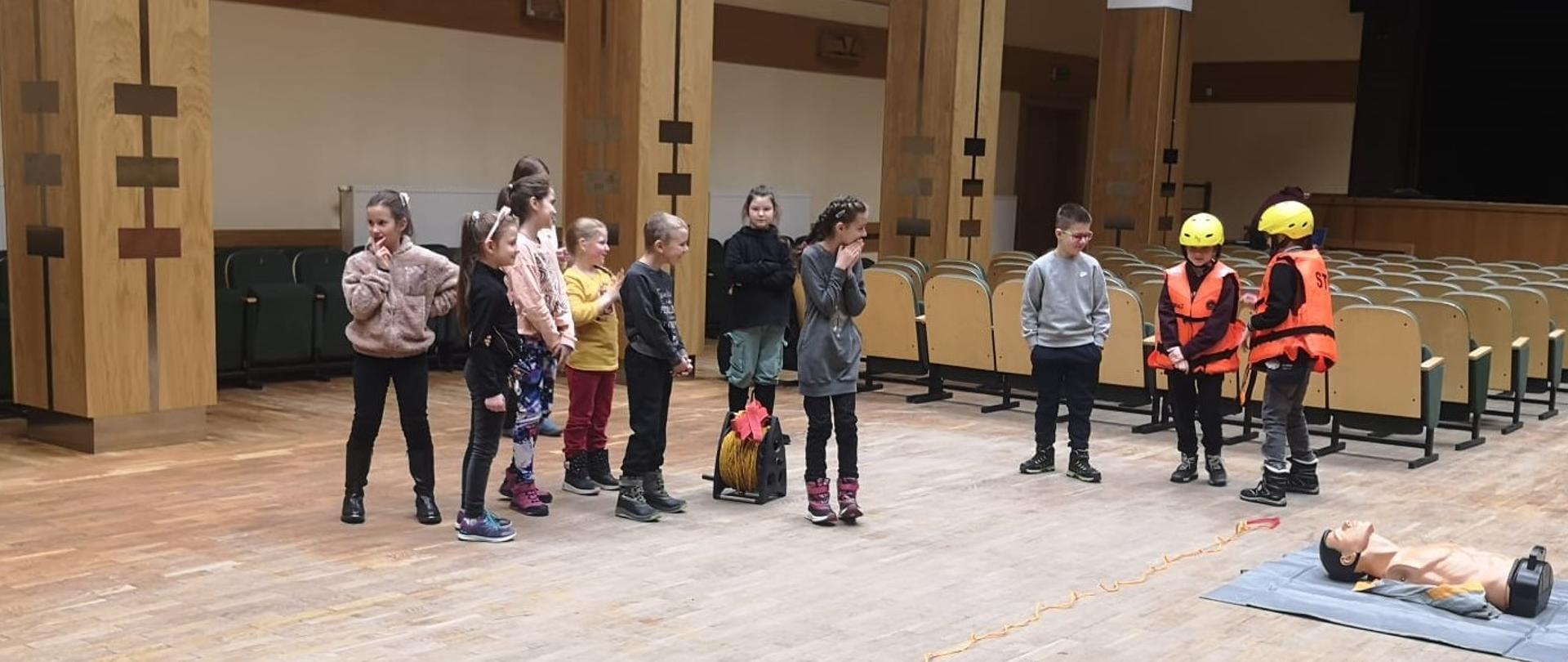 W dużym pomieszczeniu znajduje się kilkoro dzieci. Dwójka z nich ma żółte hełmy i pomarańczowe kamizelki.