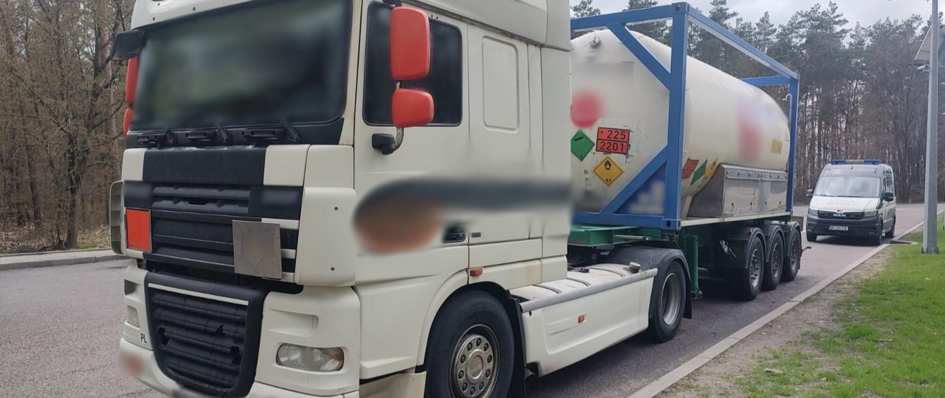 Na pierwszym planie: przód i lewy bok ciągnika siodłowego z naczepą przewożącą kontener – cysternę ADR. W tle: przód i lewy bok oznakowanego furgonu lubelskiej Inspekcji Transportu Drogowego.
