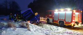 Zdjęcie przedstawia przewrócony samochód dostawczy. W tle znajduje się zaśnieżony las. Obok samochodu stoją strażacy. Jeden z nich przykłada do samochodu drabinę.