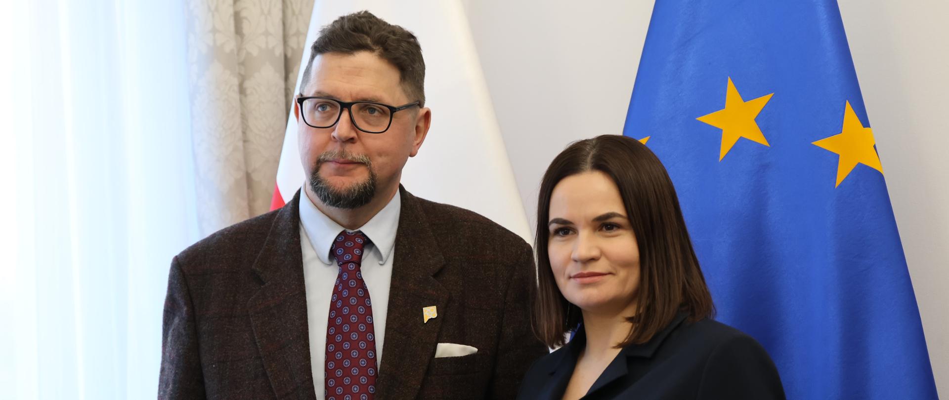 Wiceminister Szeptycki stoi obok kobiety w czarnej sukience, za nimi flagi Polski i UE.