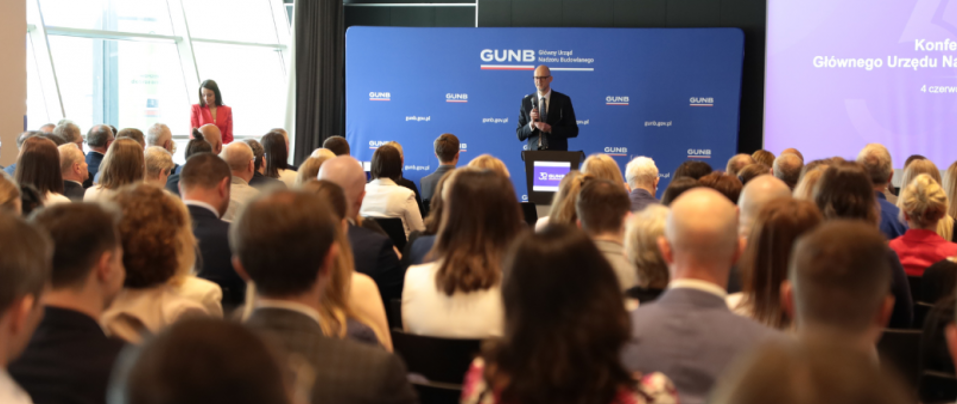 Uczestnicy konferencji szkoleniowo-jubileuszowej Głównego Urzędu Nadzoru Budowlanego (GUNB) biorą udział w obradach, prezentacjach oraz wspólnych sesjach zdjęciowych. Wydarzenie odbywa się w sali konferencyjnej z materiałami promocyjnymi GUNB w tle.