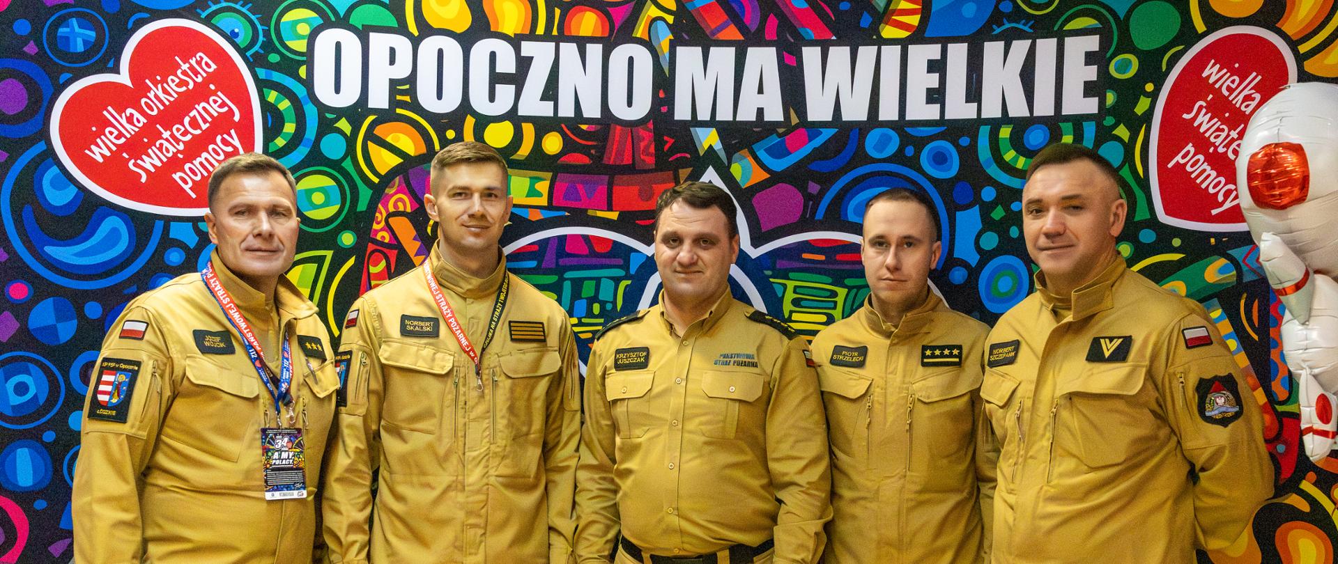 Pięciu funkcjonariuszy Państwowej Straży Pożarnej w umundurowaniu służbowym koloru piaskowego, pozuje do zdjęcia na tle ścianki 34 Finału Wielkiej Orkiestry Świątecznej Pomocy