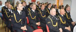 Świetlica komendy. Na krzesłach siedza uczestnicy uroczystej zbiórki. Sa to funkcjonariusze komendy. Występują w umundurowaniu wyjściowym ze sznurem. W tle ściana z logo PSP.
