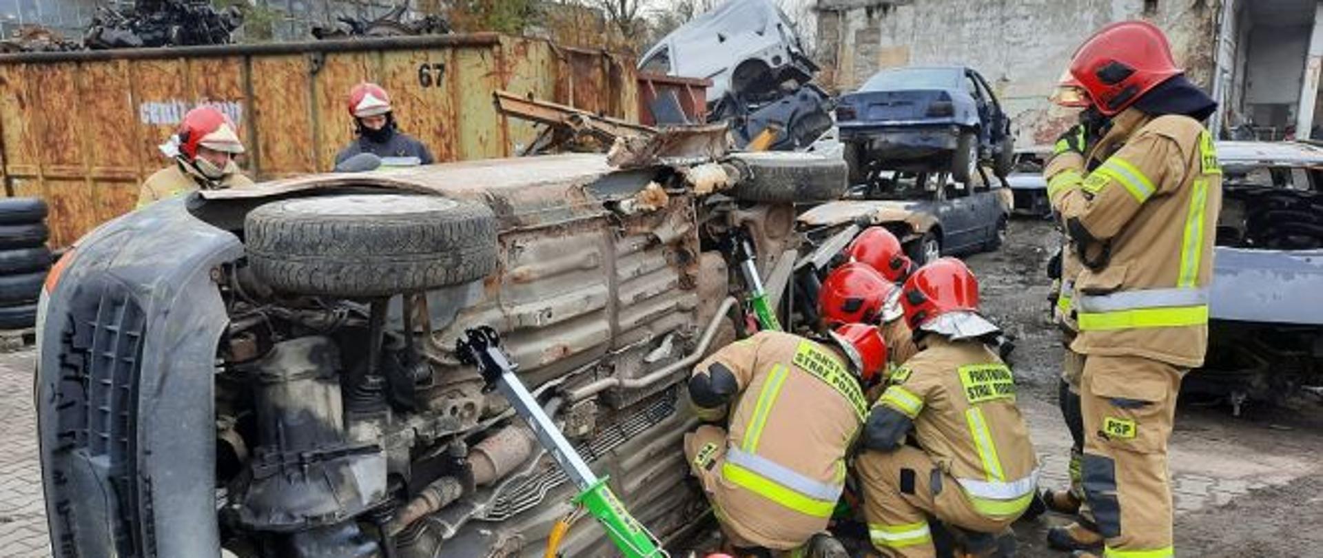 Ratownicy w trakcie ćwiczeń z ratownictwa drogowego wykonujący stabilizację pojazdu przewróconego na bok. Na kostce brukowej leży przewrócony na bok samochód osobowy. Sześciu ratowników prowadzi działania polegające na jego stabilizacji. Przednia część pojazdu została już zabezpieczona. Trzech ratowników prowadzi działania zabezpieczenia pojazdu w jego tylnej częći. Kucając zakładają podpory. W tle widać metalowe kontenery oraz budynki.