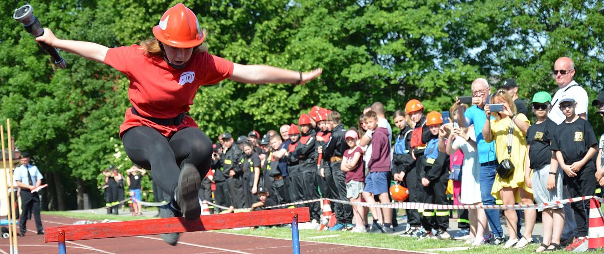 Zdjęcie przedstawia zawodniczkę podczas skoku przez płotek, w tle ludzie
