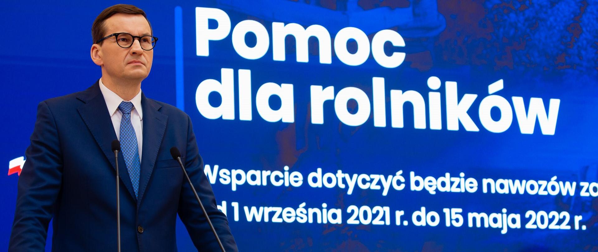 Na zdjęciu premier RP podczas konferencji prasowej. Premier stoi za mównicą, w tle duży ekran, na którym wyświetlona plansza informacyjna. 