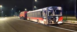 Spalony autobus na poboczu drogi holowany przez samochód ratownictwa technicznego.