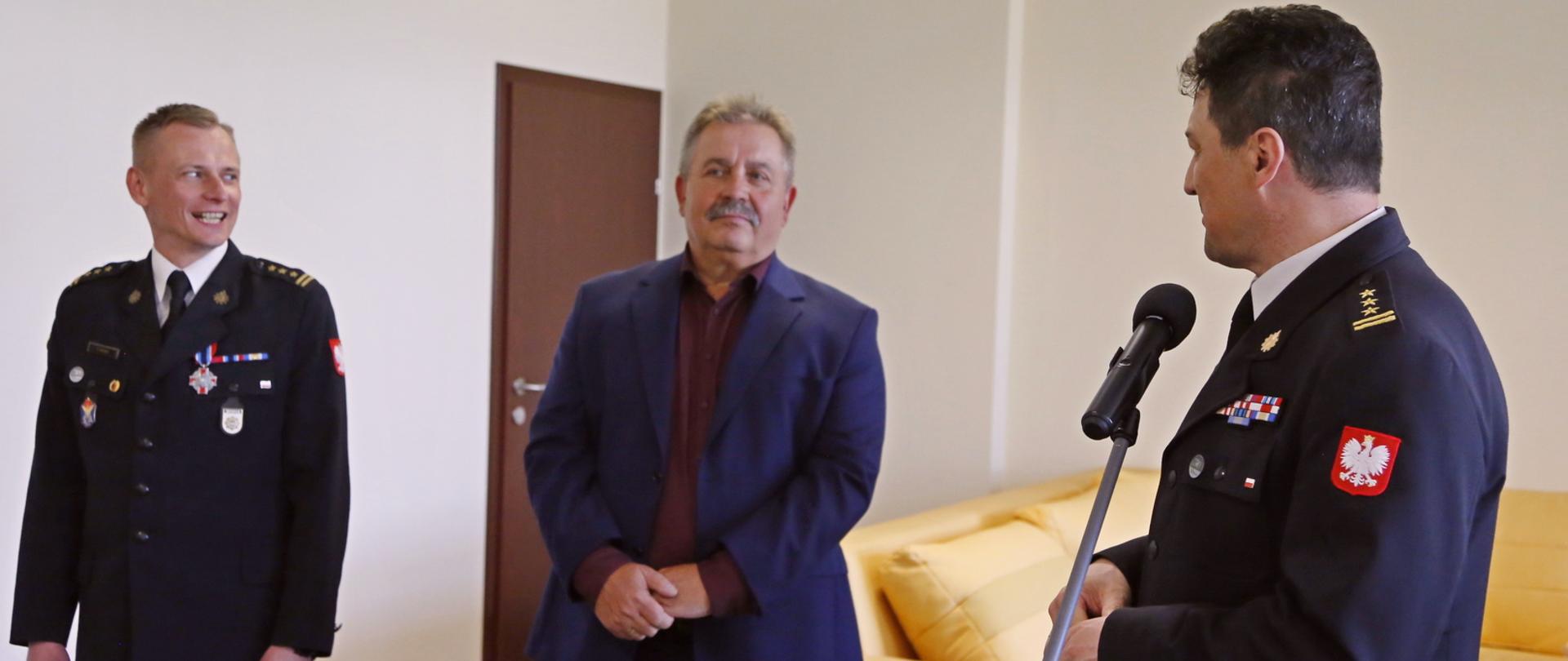Od lewej strony stojący Zastępca Komendanta Centralnej Szkoły PSP, mężczyzna oraz Komendant Centralnej Szkoły PSP przemawiający do mikrofonu