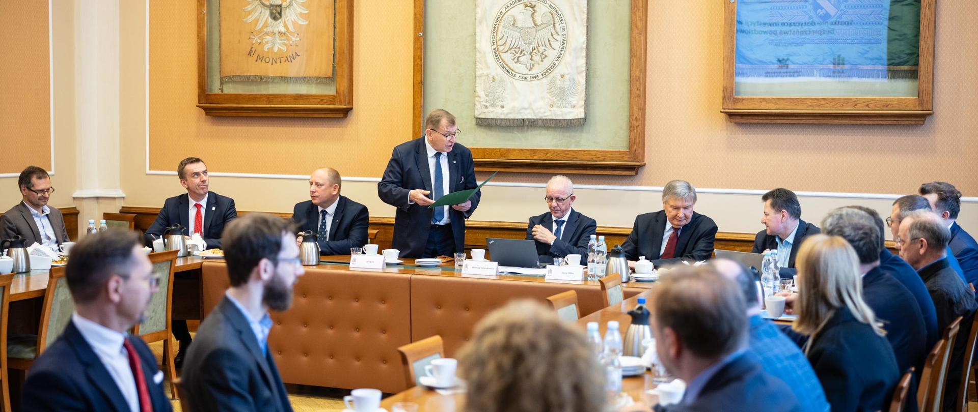 Zdjęcie przedstawia spotkanie przy dużym, prostokątnym stole w sali konferencyjnej. Uczestniczy w nim kilkanaście osób W tle widoczne są obrazy i elegancka, klasyczna aranżacja wnętrza