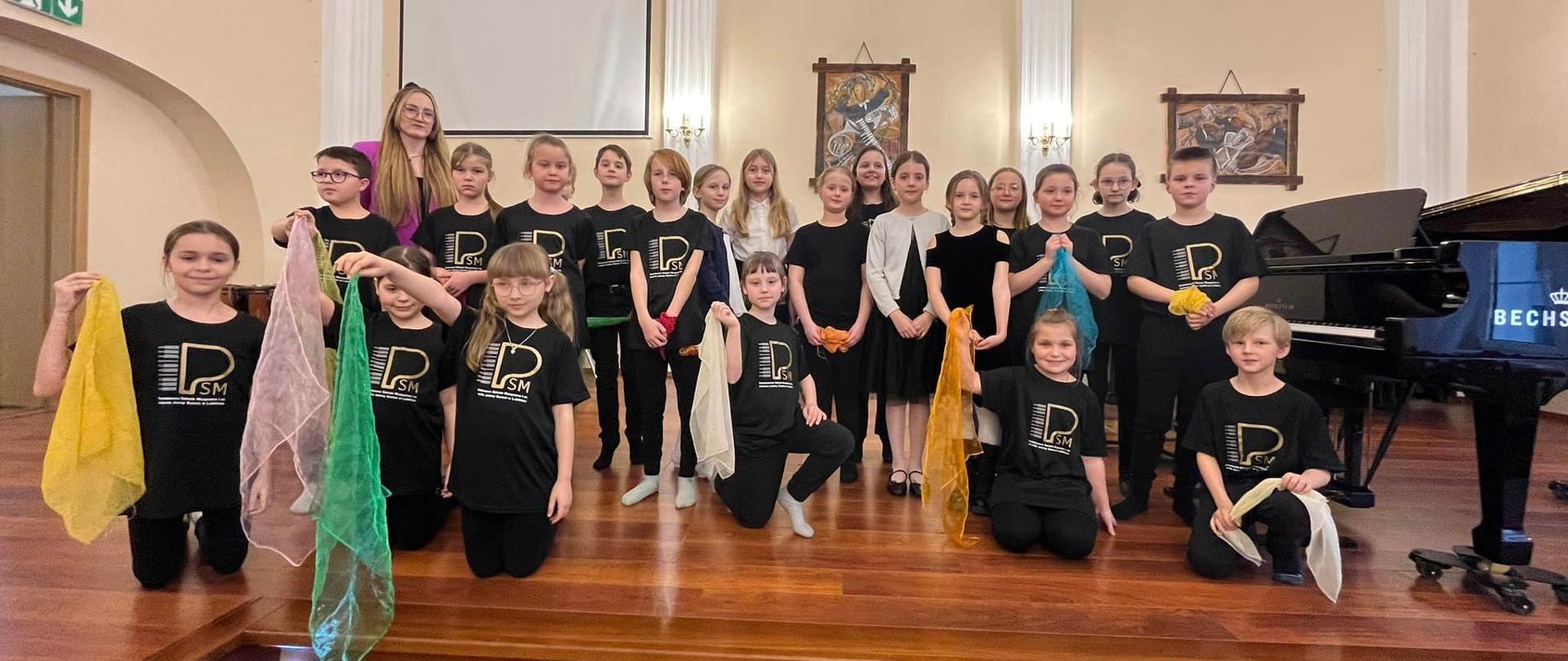 Uczniowie klasy III/6 na scenie auli podczas popisu w czarnych koszulkach z logo szkoły i kolorowymi chustami w rękach wraz z nauczycielką