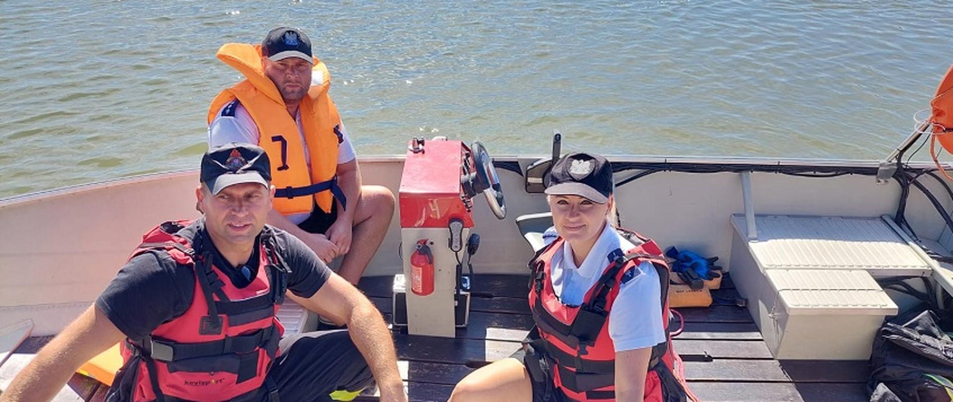 Obraz przedstawia strażaka i dwoje policjantów na łodzi ratowniczej na zbiorniku Słup. Pora dzienna.