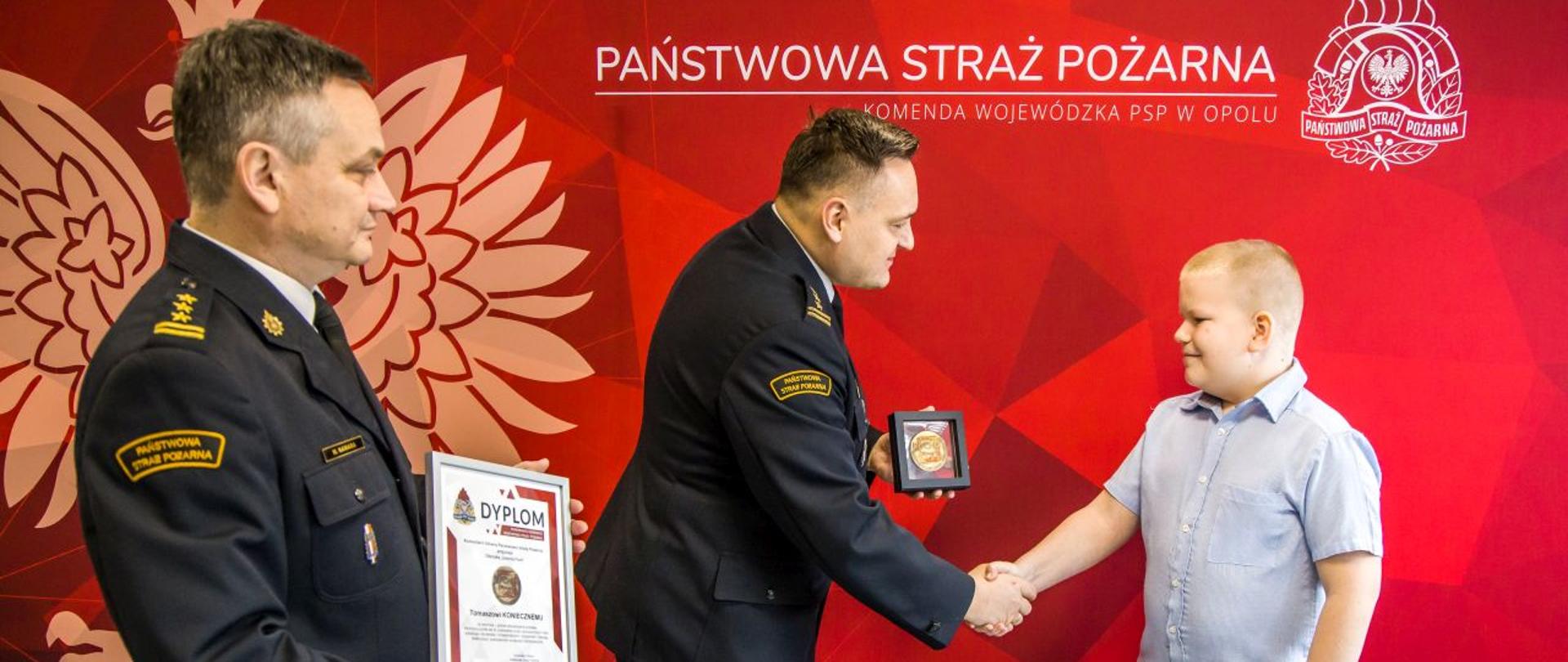 Opolski Komendant Wojewódzki st. bryg. Krzysztof Kędryk w obecności Zastępcy OKW PSP w Opolu st. bryg. Wojciecha Nawary wręcza Tomaszowi Koniecznemu Odznakę Komendanta Głównego PSP „Iuvenis Forti” za swoją bohaterską postawę.
W tle widoczna plansza koloru czerwonego z białym napisem Państwowa Straż Pożarna i Komenda Wojewódzka PSP w Opolu.