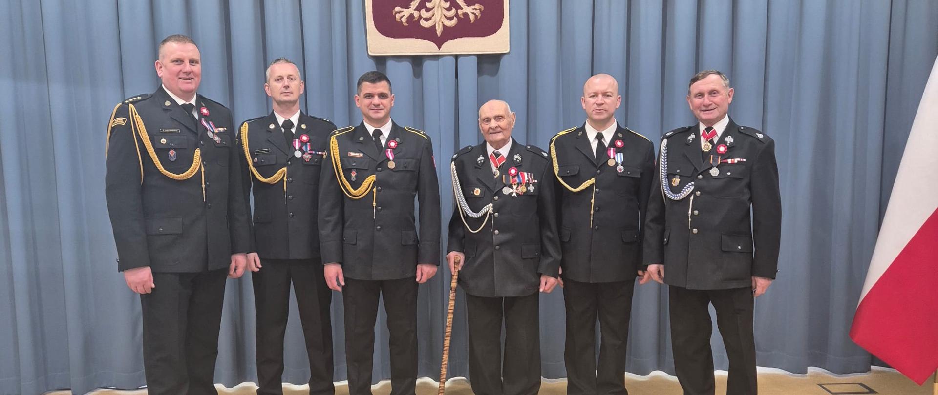 Grupa odznaczonych strażaków PSP i druhów OSP z powiatu przasnyskiego pozuje do wspólnego zdjęcia na tle godła państwowego.