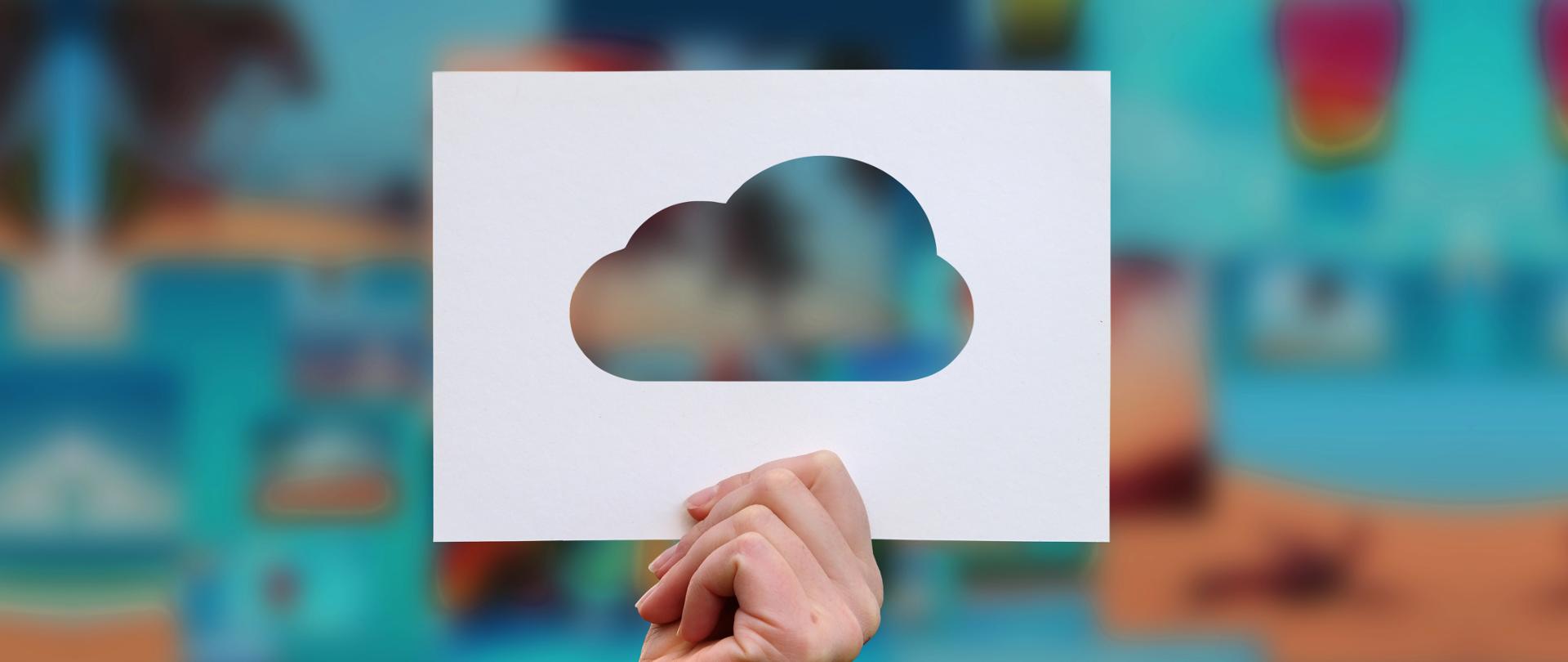 Damska dłoń trzymająca białą kartkę, na środku której wycięty jest kształt chmury, kolorowe, rozmazane tło.