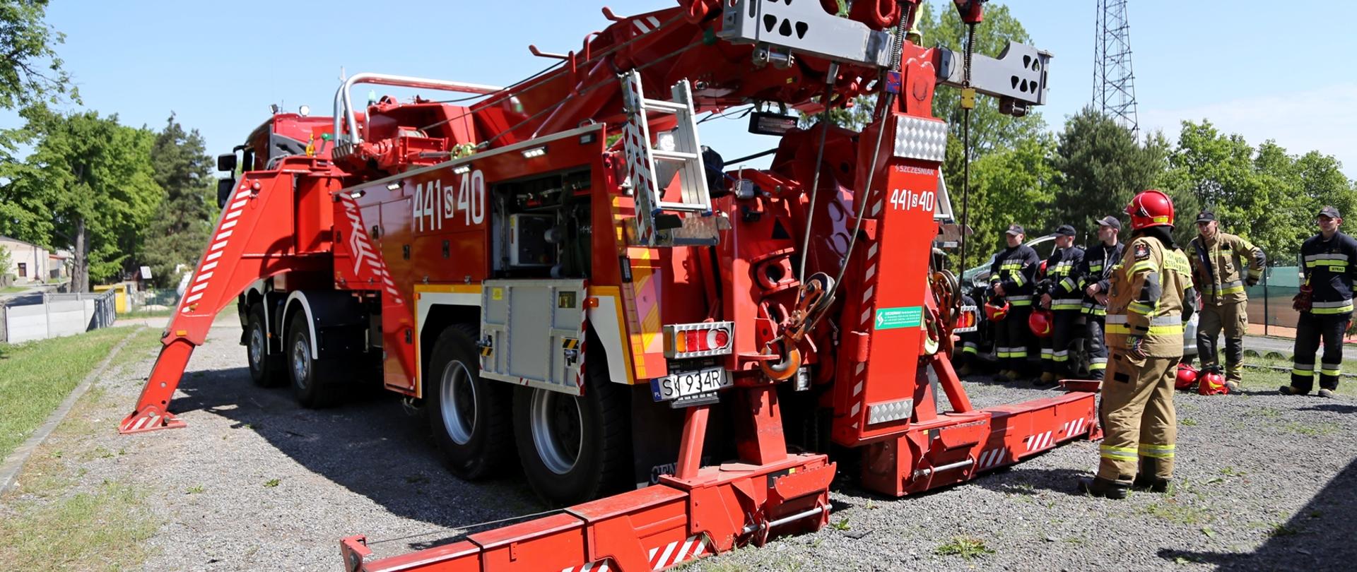 Widok z tyłu na samochód pożarniczy SCRT „Rotator”, który ma rozstawione podpory. W tle strażacy obserwujący pokaz