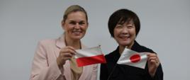 Dwie kobiety pozują do zdjęcia. Jedna trzyma flagę Polski druga Japonii.