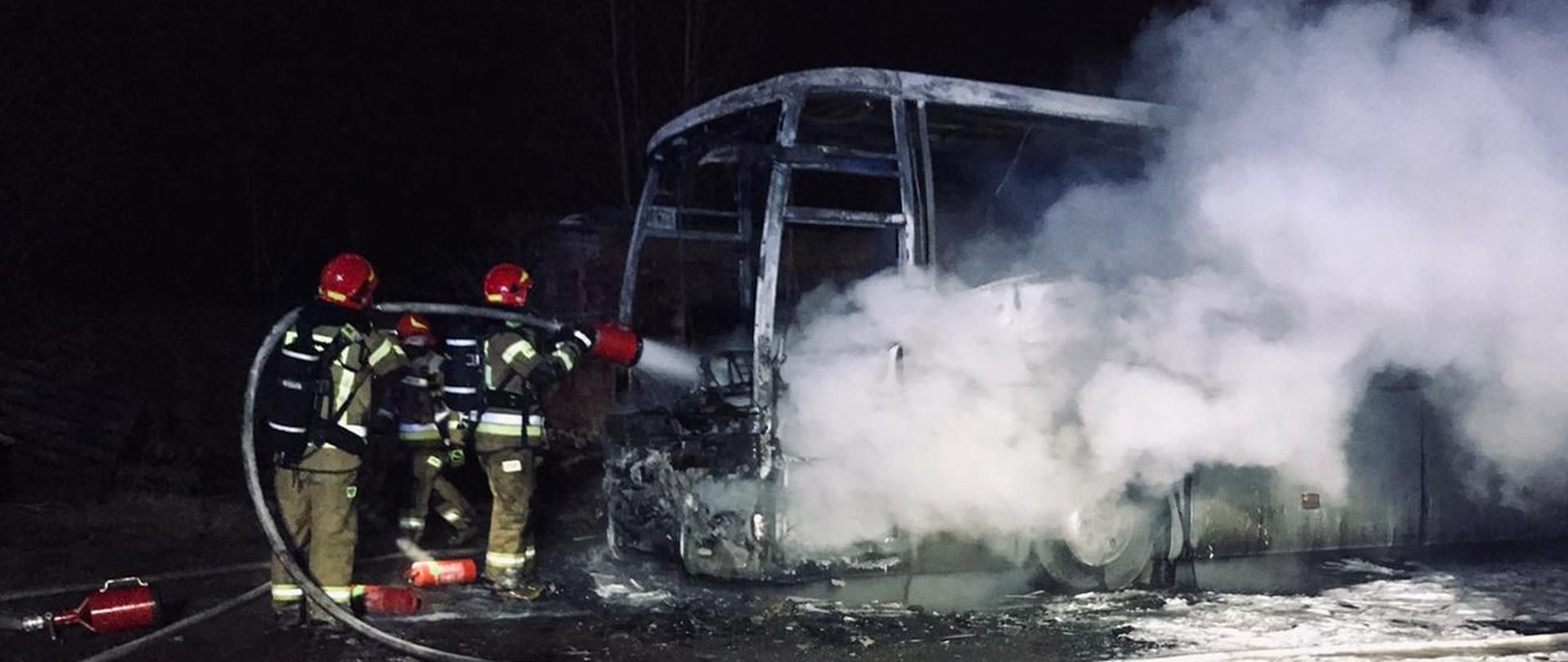 Zdjęcie przedstawia na ciemnym tle trzech strażaków ubranych w ubrania specjalne gaszących palący się autobus za pomocą wytwornicy pianowej. Strażacy stoją po lewej stronie, z prawej strony z palącego się pojazdu wydobywa się gęsty biały dym