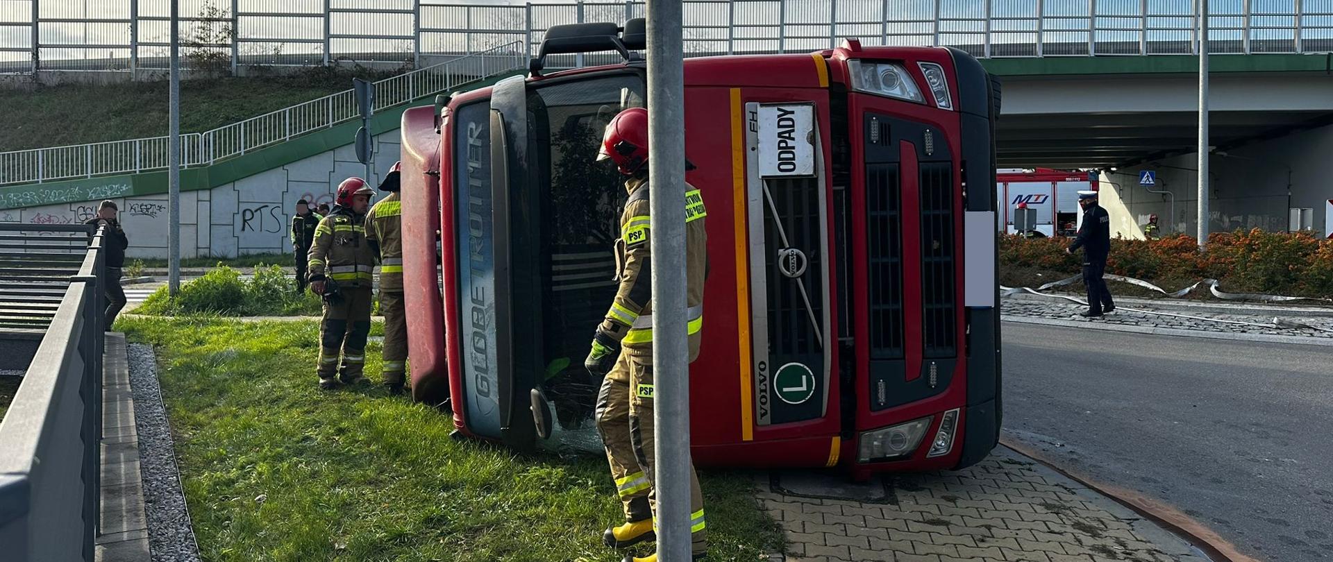 Samochód ciężarowy leży na boku, obok niego przechodzi strażak i stoi kilku innych strażaków.