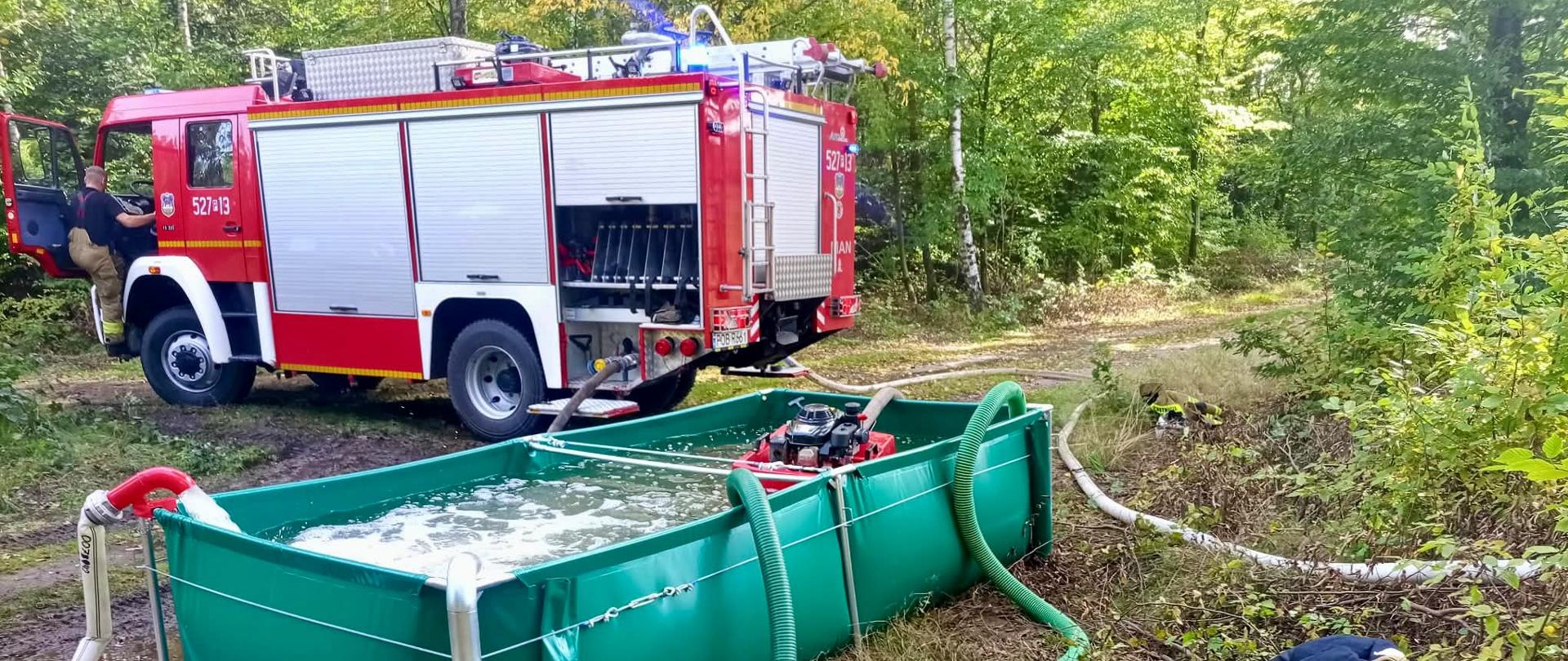 Na leśnej drodze stoi czerwony wóz strażacki, a przed nim znajduje się duży zielony zbiornik na wodę wypełniony wodą. Zbiornik jest połączony licznymi wężami z pompami spalinowymi i wozem strażackim. Po prawej stronie na ziemi leżą elementy wyposażenia strażackiego, w tym hełm i ubranie ochronne. Wokół znajduje się gęsty, zielony las.