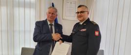 Wójt gminy Nowe i komendant wojewódzki PSP stoją przy stole, po podpisaniu umowy i podają sobie dłonie. W reku Wójta podpisany dokument. W tle wystrój gabinetu komendanta.