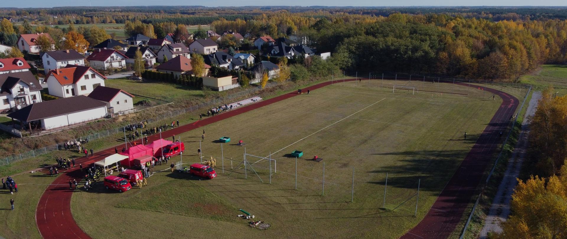 Zdjęcie przedstawia widok z lotu ptaka na boisko sportowe otoczone bieżnią oraz tereny mieszkalne i leśne w jesiennej scenerii.
Centralny punkt: Duże, trawiaste boisko (prawdopodobnie do piłki nożnej) w kształcie owalu, z widocznymi gdzie nie gdzie elementami wyposażenia sportowego (bramki, pachołki).
Bieżnia: Wokół boiska biegnie czerwono-brązowa, syntetyczna bieżnia o charakterystycznym, podłużnym kształcie.
Wydarzenie/Ludzie: Na fragmencie boiska wzdłuż lewej krawędzi (na zakręcie) widoczna jest grupa ludzi, zawodnicy OSP oraz czerwone pojazdy straży pożarnej i rozstawiony czerwony namiot/konstrukcja, co sugeruje, że na obiekcie odbywa się jakieś lokalne wydarzenie lub zawody strażackie.
