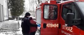Zdjęcie przedstawia samochód strażacki na tle wejścia do budynku Niepublicznego Zakładu Opieki Zdrowotnej w Dwikozach. Na zdjęciu widać jak strażak pomaga wyjść osobie starszej z samochodu strażackiego. 