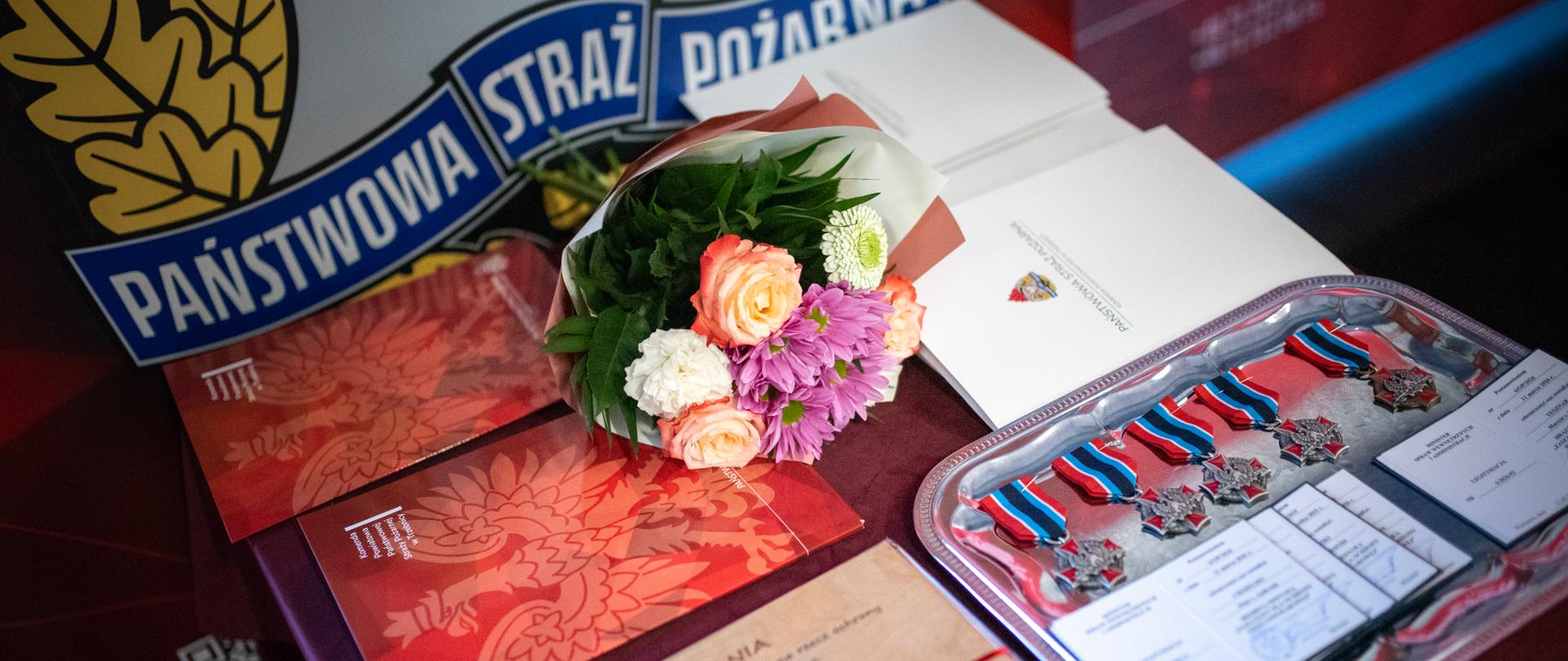 Na stoliku leżą odznaczenia strażackie, dyplomy i bukiet kwiatów przygotowane na uroczystość Państwowej Straży Pożarnej.