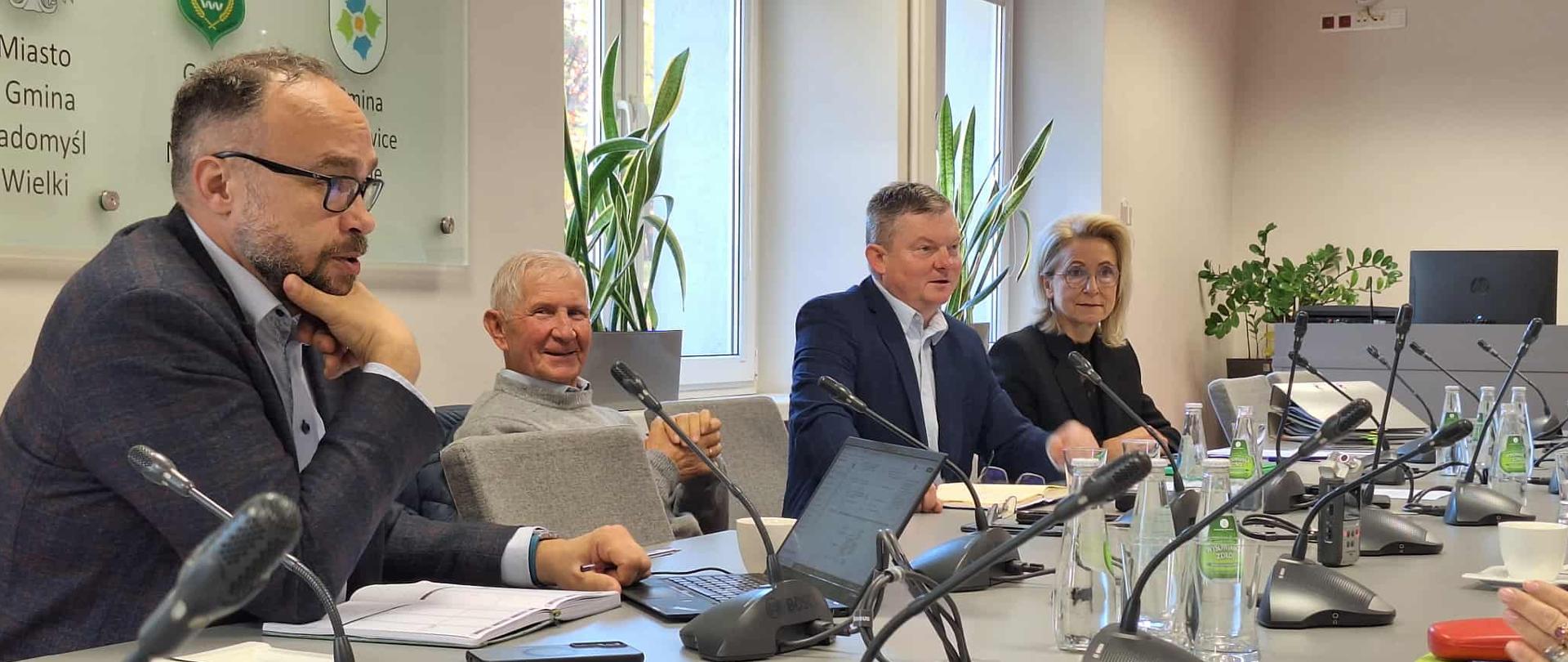 Grupa ludzi siedząca w jasnym pomieszczeniu przy stole na którym są dokumenty. 