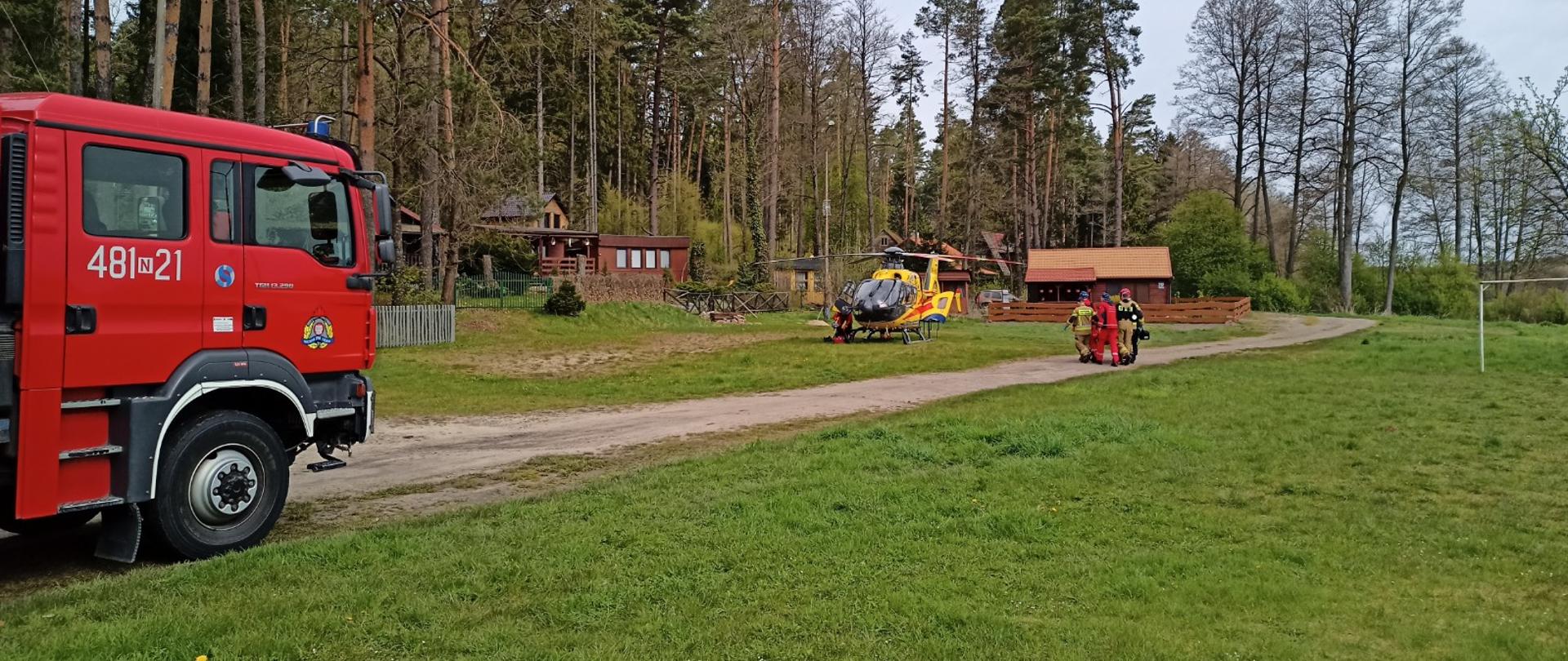 Zdjęcie przedstawia wóz strażacki, smigłowiec LPR oraz strażaków i ratowników naszących poszkodowanego z ROSC do smigłowca.
