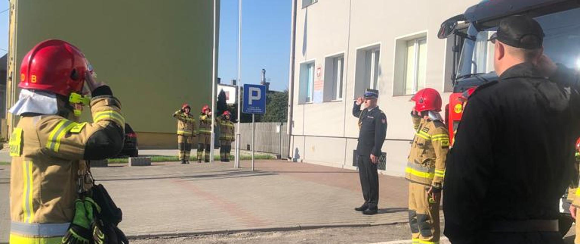 zdjęcie zrobione w dzień przed budynkiem Komendy Powiatowej PSP w Sępólnie Krajeńskim. Przedstawia strażaków podczas uroczystej zmiany służby z okazji podniesienia flagi państwowej. Strażacy w umundurowaniu bojowym stoją na przeciwko siebie - dwie zmiany służbowe. Od tył widoczny zastępca dowódcy JRG. W oddali przy maszcie stoi poczet flagowy - 3 strażaków w umundurowaniu bojowym, którzy wciągają flagę na maszt. Strażacy oddają honory. 