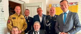 Grupa osób, w tym oficerowie i wicewojewoda, pozuje do zdjęcia z uhonorowanym strażakiem siedzącym na krześle. Strażak trzyma w rękach godło Polski otrzymane podczas uroczystości.