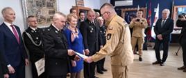 Zastępca komendanta głównego ubrany w służbowy strój w kolorze piaskowym wręcza strażakowi ochotnikowi promese 