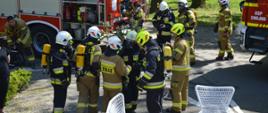 Ćwiczenia w Pakosławiu. Na tle wozów strażackich i parku znajdują się strażacy uczestniczący w ćwiczeniach. Mają białe i żółte hełmy. Część z nich wyposażona jest w aparaty oddechowe.