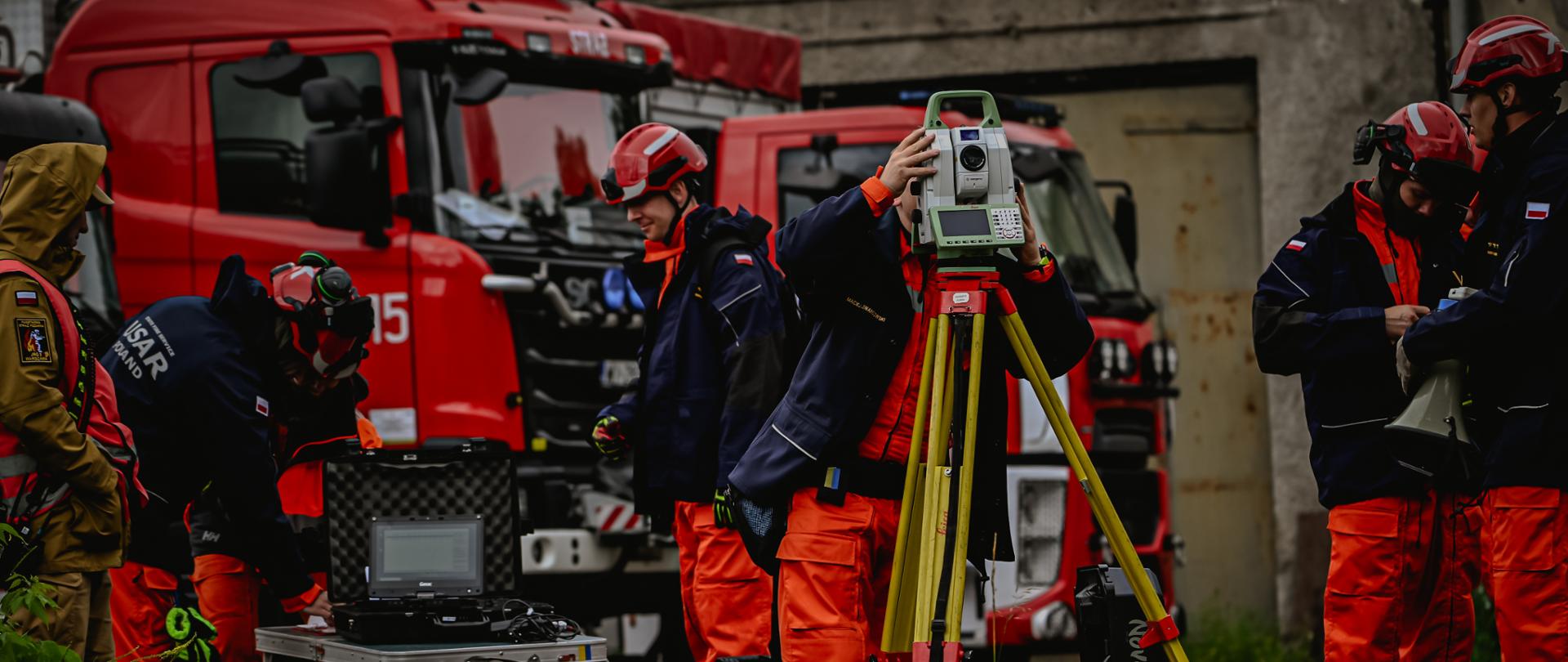 Na pierwszym planie, na kamienistym placu przed budynkiem, stoi strażak obsługujący zaawansowany instrument geodezyjny (tachometr) zamocowany na żółtym statywie. Po lewej stronie, na składanym stoliku turystycznym, rozstawiony jest polowy komputer (laptop) w czarnej walizce transportowej, przy którym pracują kolejni ratownicy. W tle zaparkowane są dwa duże, czerwone samochody ratowniczo-gaśnicze. Po prawej stronie kadru stoi grupa strażaków w ciemnych kurtkach i pomarańczowych spodniach, z których jeden trzyma biało-zielony megafon.