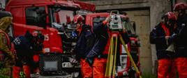 Na pierwszym planie, na kamienistym placu przed budynkiem, stoi strażak obsługujący zaawansowany instrument geodezyjny (tachometr) zamocowany na żółtym statywie. Po lewej stronie, na składanym stoliku turystycznym, rozstawiony jest polowy komputer (laptop) w czarnej walizce transportowej, przy którym pracują kolejni ratownicy. W tle zaparkowane są dwa duże, czerwone samochody ratowniczo-gaśnicze. Po prawej stronie kadru stoi grupa strażaków w ciemnych kurtkach i pomarańczowych spodniach, z których jeden trzyma biało-zielony megafon.